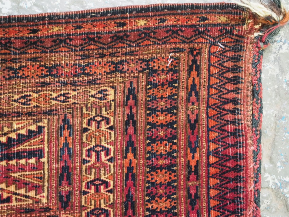 An antique woven Tekke Turkmen torba featuring intricate geometric lattice motifs in dark red, terracotta, and dark blue with a long braided fringe along the bottom edge. View X