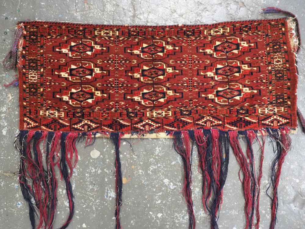 A horizontal rectangular handwoven wool Tekke Turkmen torba bag featuring nine geometric gul motifs in red, navy, and ivory, complete with a long hanging wool fringe at the base. View III