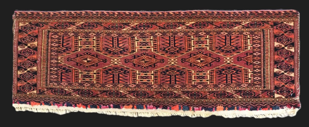 An antique rectangular hand-knotted wool Saryk Turkmen torba face displaying geometric diamond medallions in red, terracotta, and navy shades with lower fringe detail.