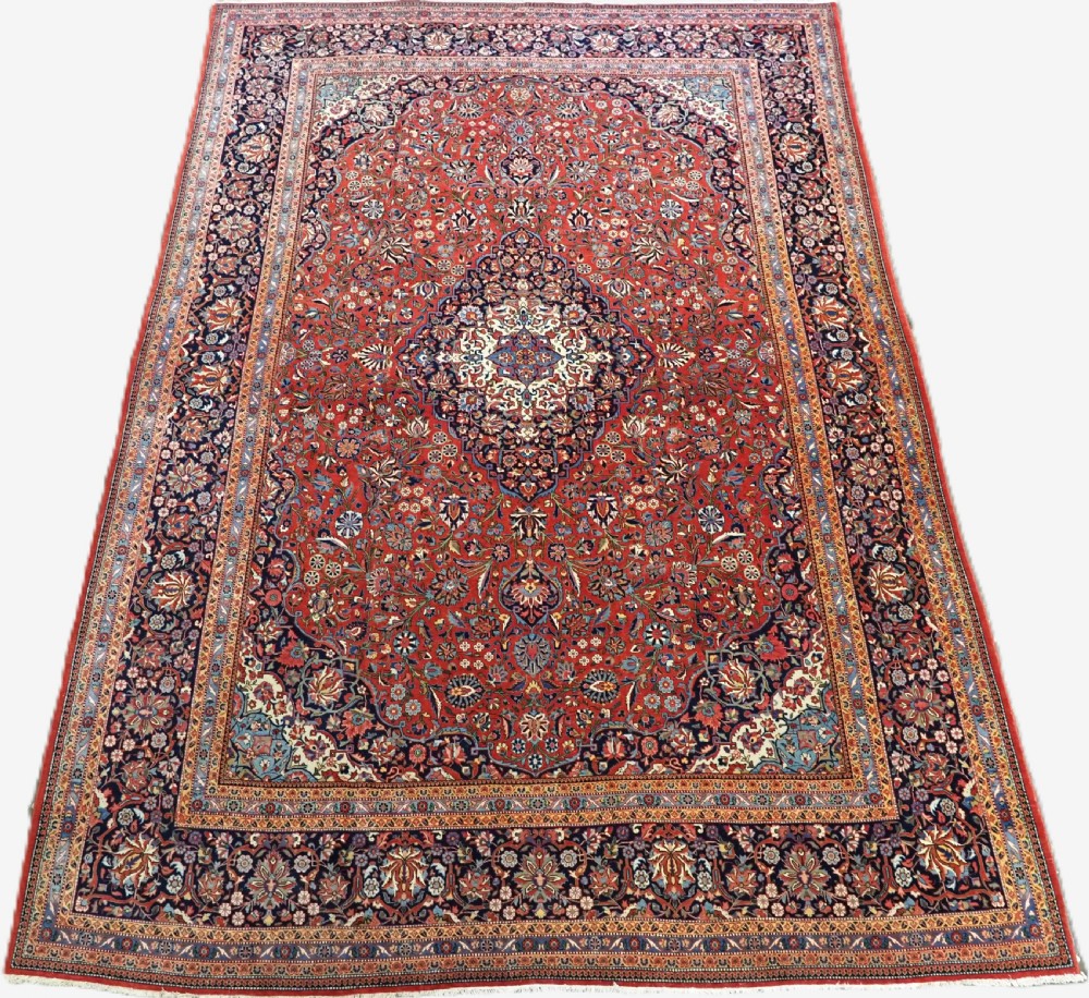A large hand-knotted antique Kashan carpet featuring a central navy and ivory medallion on a crimson field with intricate floral patterns and layered borders.