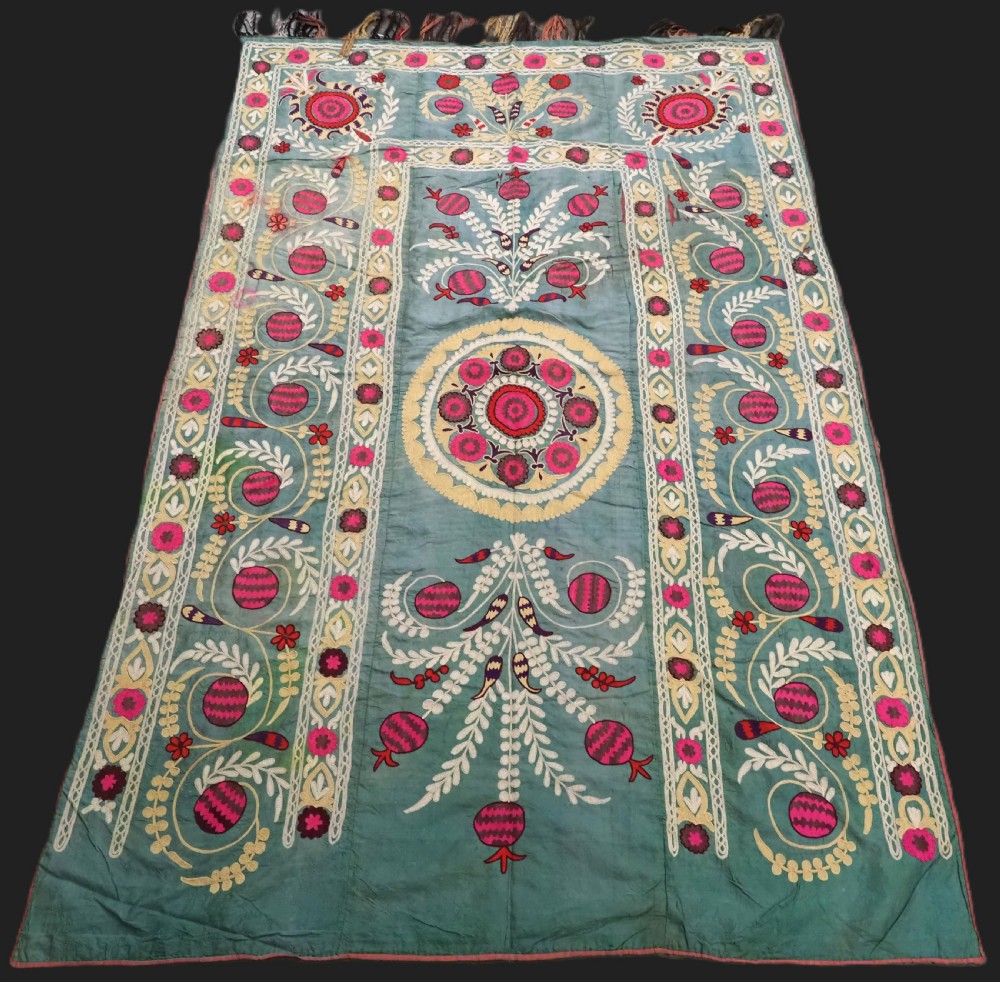 An antique teal silk suzani textile embroidered with pink and red pomegranate motifs, floral medallions, and decorative borders with top fringe.