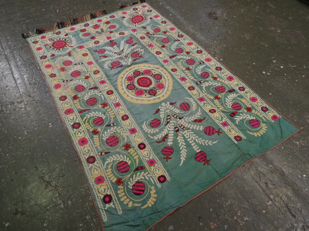 An antique teal silk suzani textile embroidered with pink and red pomegranate motifs, floral medallions, and decorative borders with top fringe. View II