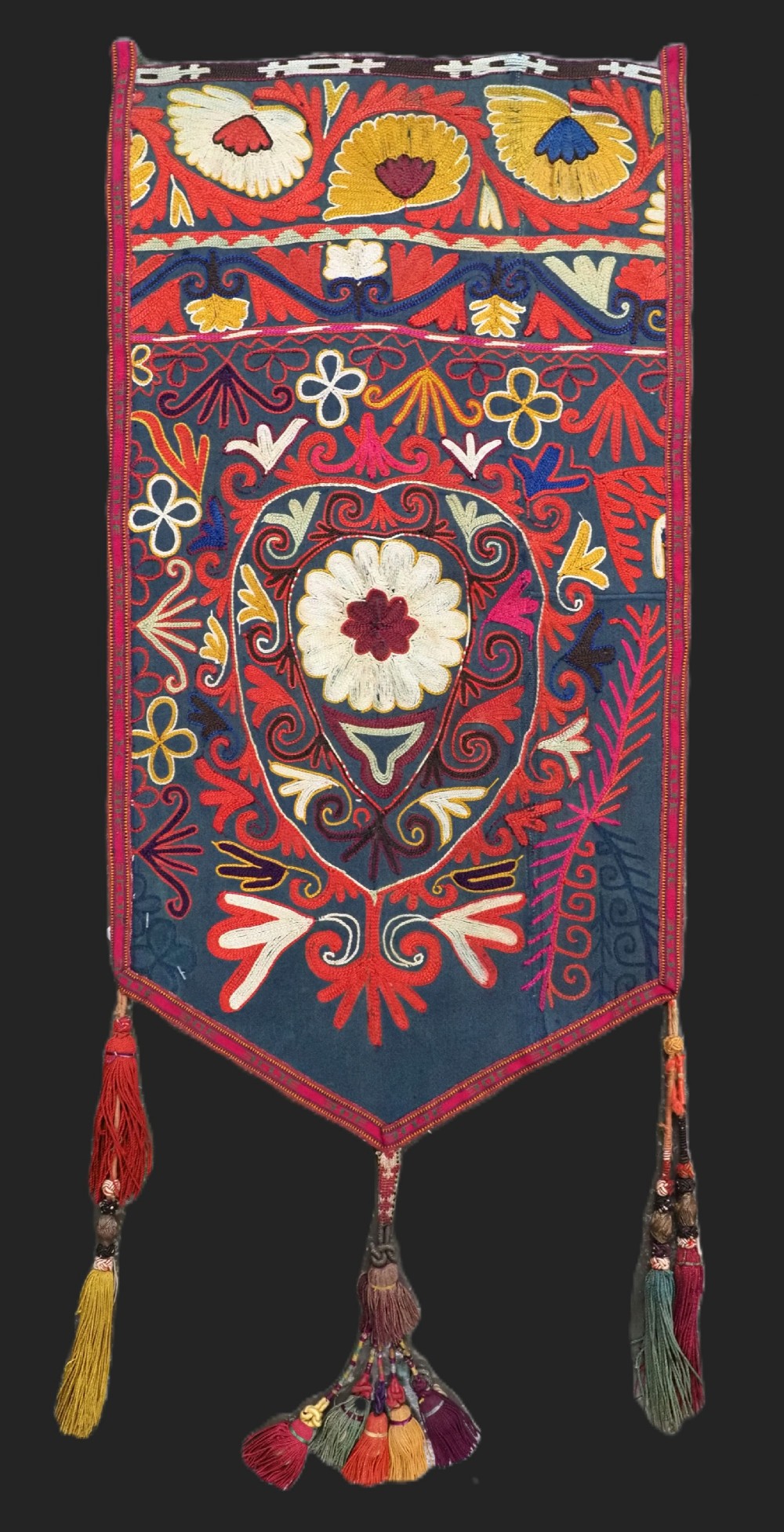 A shield-shaped Uzbek Lakai embroidered textile featuring vibrant silk floral and scroll motifs on a dark green background with long decorative tassels along the lower edge.