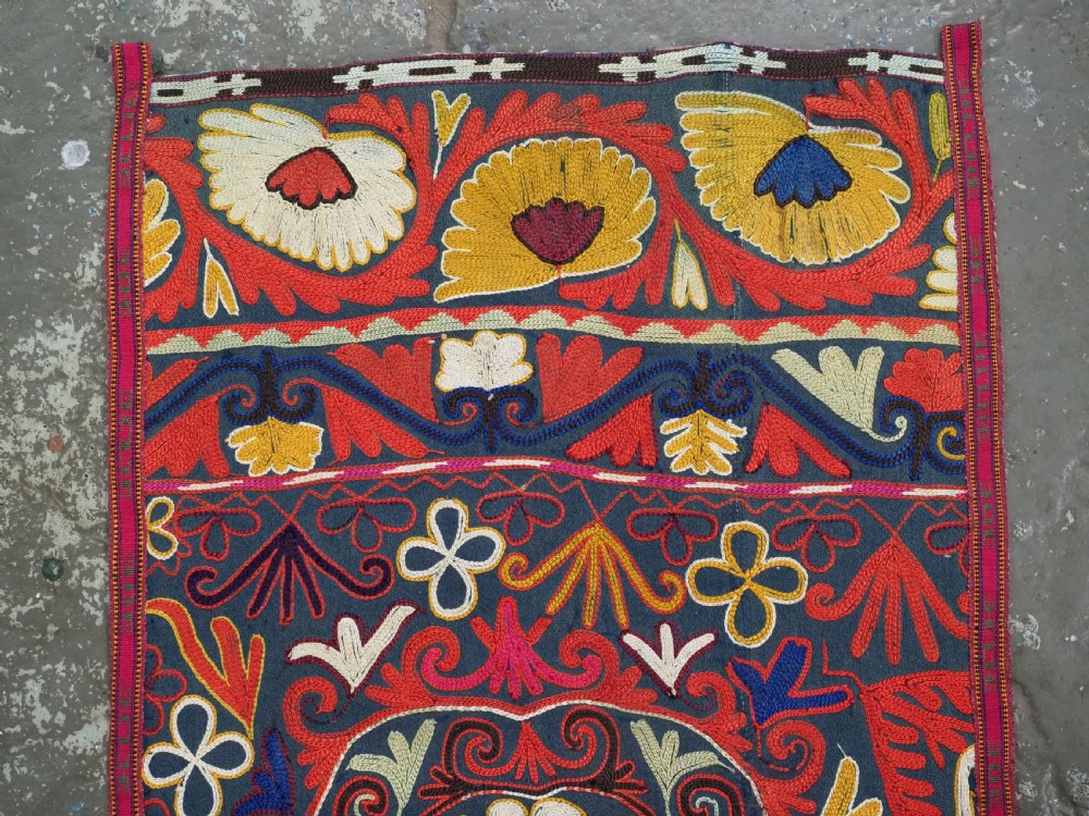 A shield-shaped Uzbek Lakai embroidered textile featuring vibrant silk floral and scroll motifs on a dark green background with long decorative tassels along the lower edge. View II