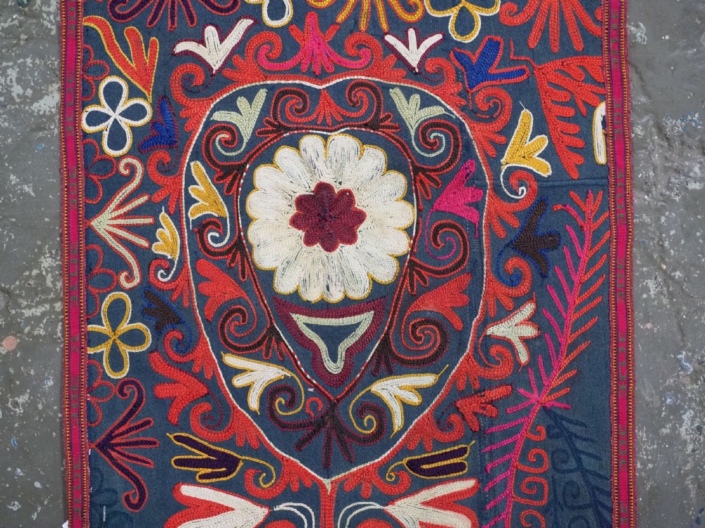 A shield-shaped Uzbek Lakai embroidered textile featuring vibrant silk floral and scroll motifs on a dark green background with long decorative tassels along the lower edge. View III