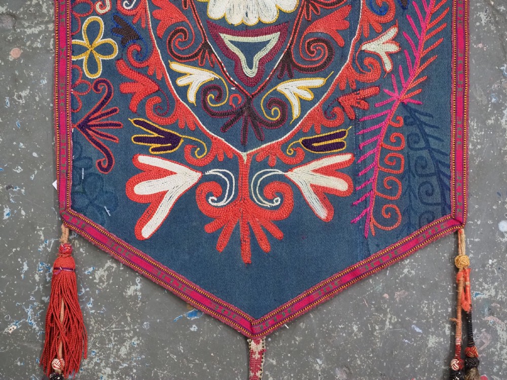 A shield-shaped Uzbek Lakai embroidered textile featuring vibrant silk floral and scroll motifs on a dark green background with long decorative tassels along the lower edge. View IV