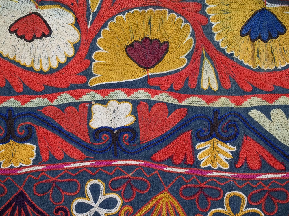 A shield-shaped Uzbek Lakai embroidered textile featuring vibrant silk floral and scroll motifs on a dark green background with long decorative tassels along the lower edge. View V