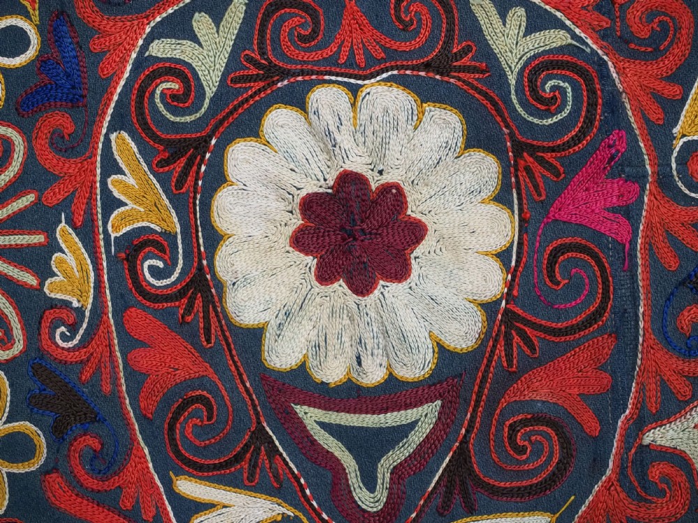 A shield-shaped Uzbek Lakai embroidered textile featuring vibrant silk floral and scroll motifs on a dark green background with long decorative tassels along the lower edge. View VI