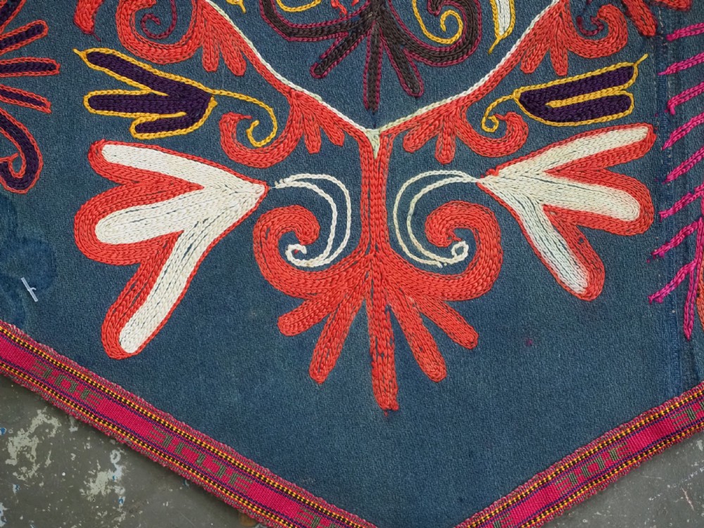 A shield-shaped Uzbek Lakai embroidered textile featuring vibrant silk floral and scroll motifs on a dark green background with long decorative tassels along the lower edge. View VII