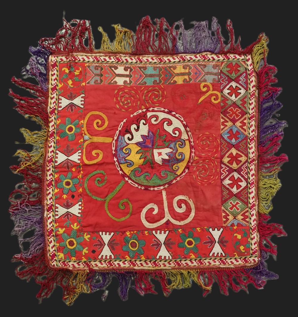 A square antique Uzbek Lakai embroidered textile featuring red cloth decorated with bright silk spiral motifs, geometric borders, and a multi-coloured fringe.