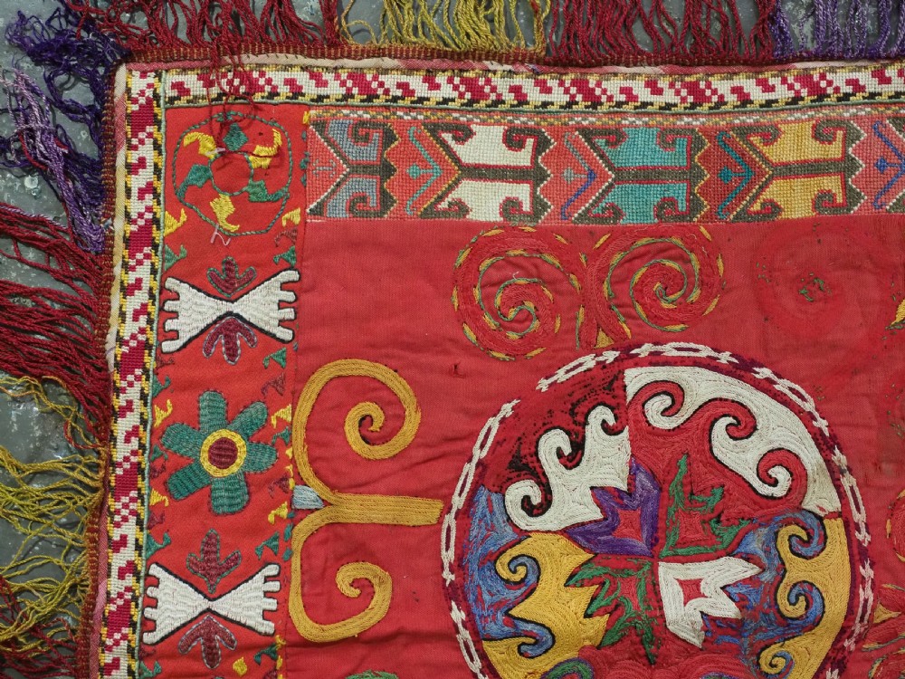 A square antique Uzbek Lakai embroidered textile featuring red cloth decorated with bright silk spiral motifs, geometric borders, and a multi-coloured fringe. View II
