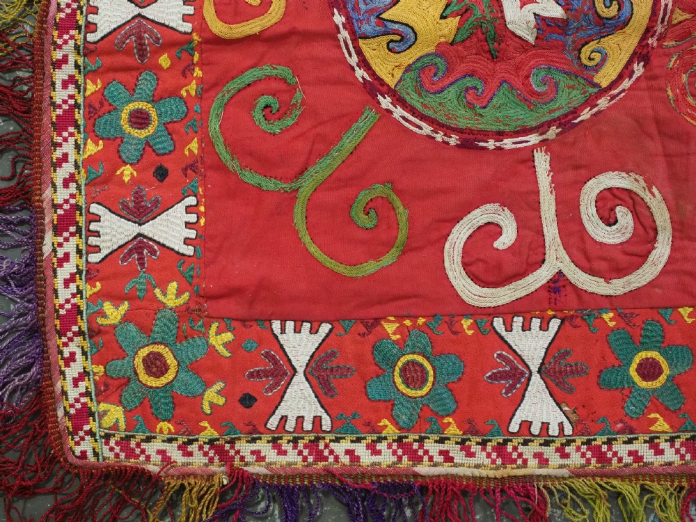 A square antique Uzbek Lakai embroidered textile featuring red cloth decorated with bright silk spiral motifs, geometric borders, and a multi-coloured fringe. View III