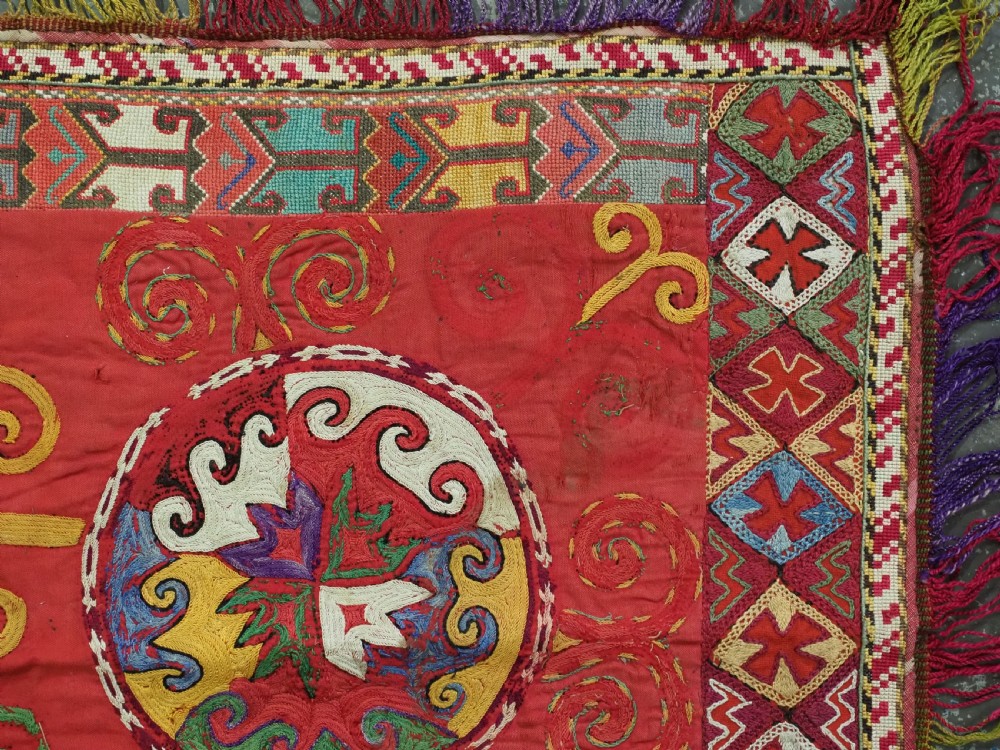 A square antique Uzbek Lakai embroidered textile featuring red cloth decorated with bright silk spiral motifs, geometric borders, and a multi-coloured fringe. View IV