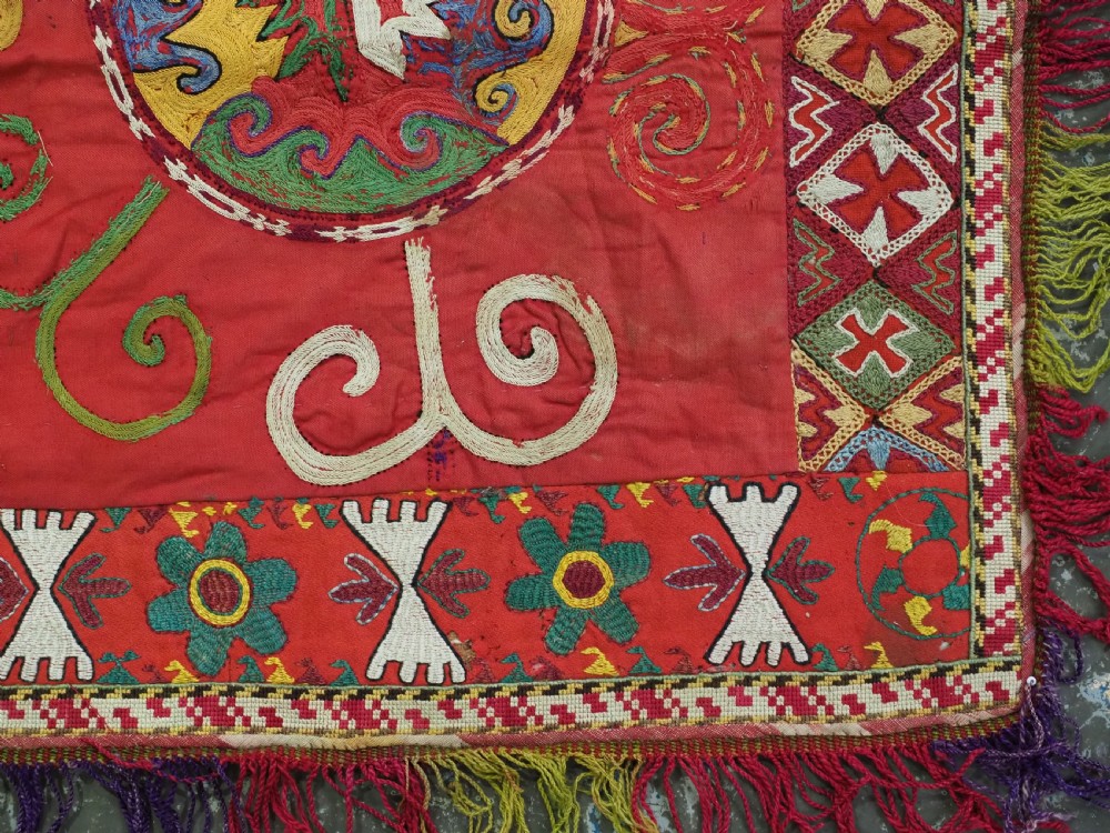 A square antique Uzbek Lakai embroidered textile featuring red cloth decorated with bright silk spiral motifs, geometric borders, and a multi-coloured fringe. View V