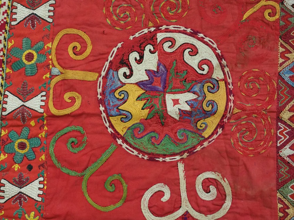 A square antique Uzbek Lakai embroidered textile featuring red cloth decorated with bright silk spiral motifs, geometric borders, and a multi-coloured fringe. View VII