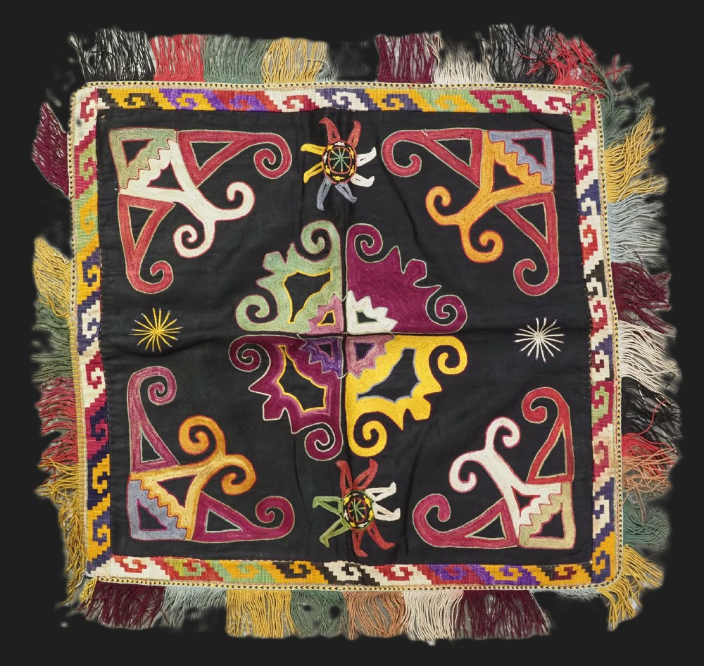A square black textile panel featuring multicoloured silk embroidery with scroll and geometric motifs bordered by a fringe edge.