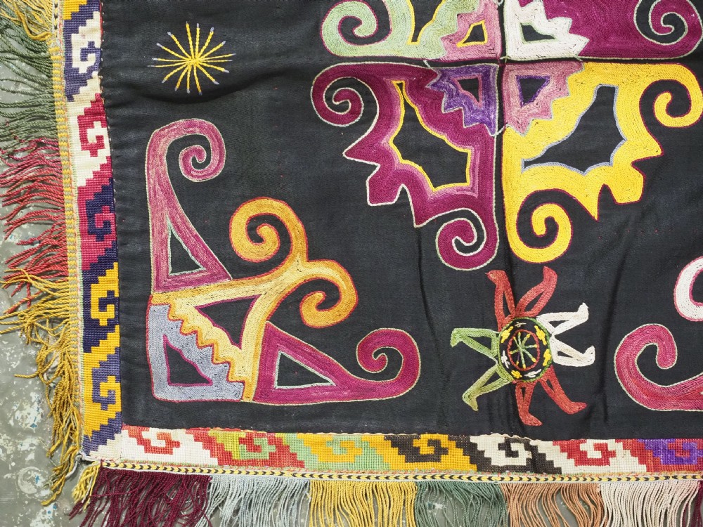 A square black textile panel featuring multicoloured silk embroidery with scroll and geometric motifs bordered by a fringe edge. View III