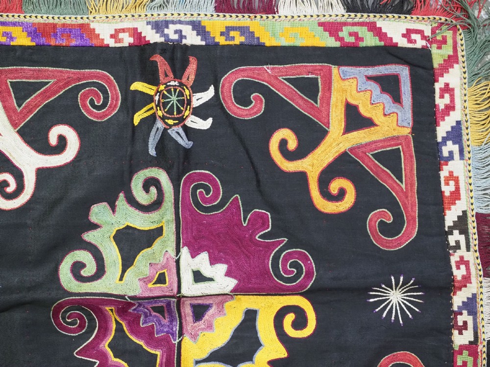 A square black textile panel featuring multicoloured silk embroidery with scroll and geometric motifs bordered by a fringe edge. View IV