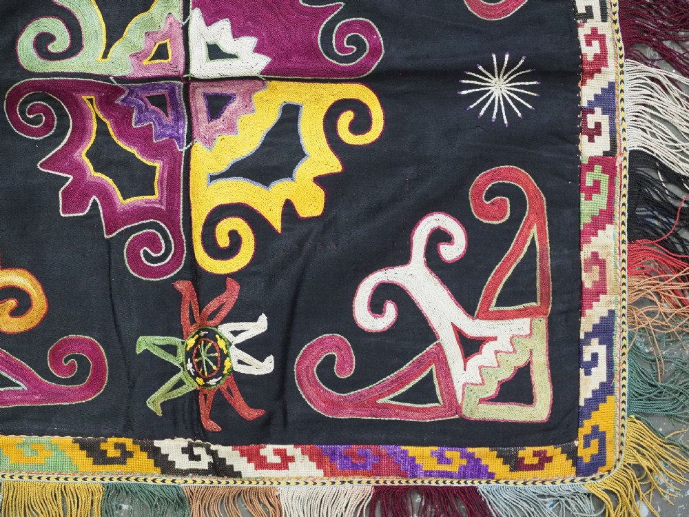 A square black textile panel featuring multicoloured silk embroidery with scroll and geometric motifs bordered by a fringe edge. View V