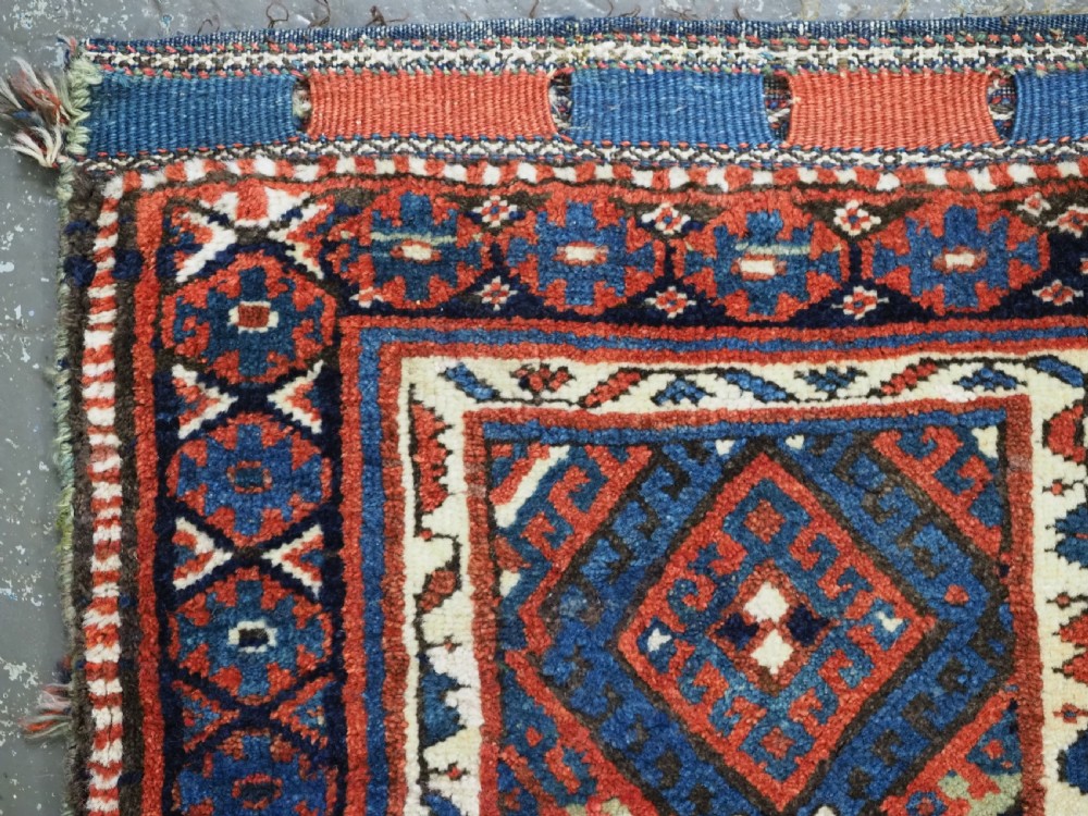 A hand-woven antique Kurdish Jaf bag featuring geometric diamond latch-hook motifs in red, blue, and cream wool with woven loops along the top edge. View III