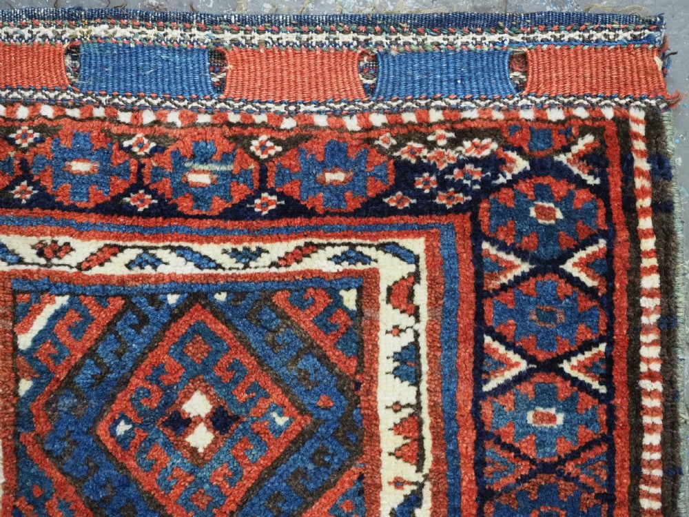 A hand-woven antique Kurdish Jaf bag featuring geometric diamond latch-hook motifs in red, blue, and cream wool with woven loops along the top edge. View V