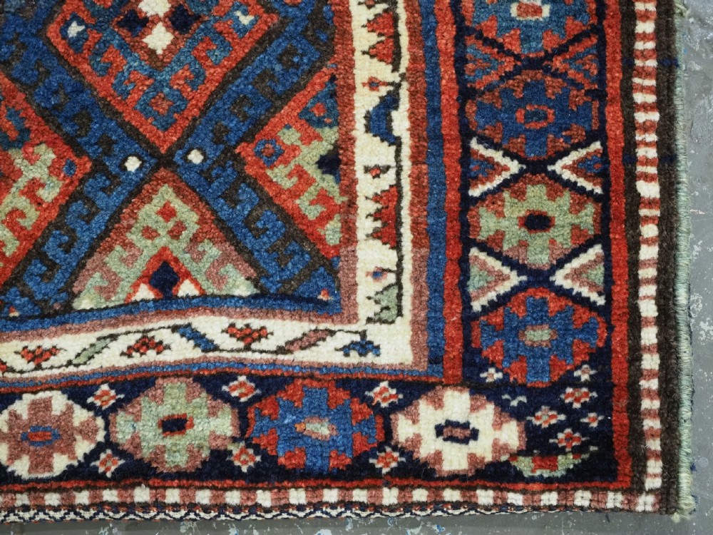 A hand-woven antique Kurdish Jaf bag featuring geometric diamond latch-hook motifs in red, blue, and cream wool with woven loops along the top edge. View VI