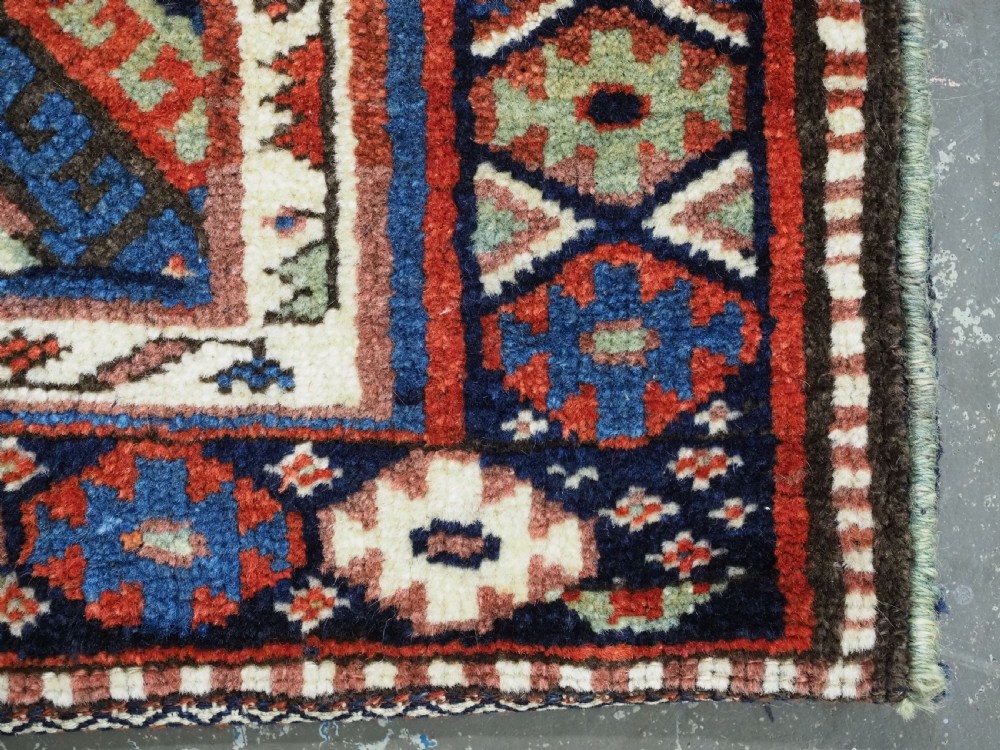 A hand-woven antique Kurdish Jaf bag featuring geometric diamond latch-hook motifs in red, blue, and cream wool with woven loops along the top edge. View X