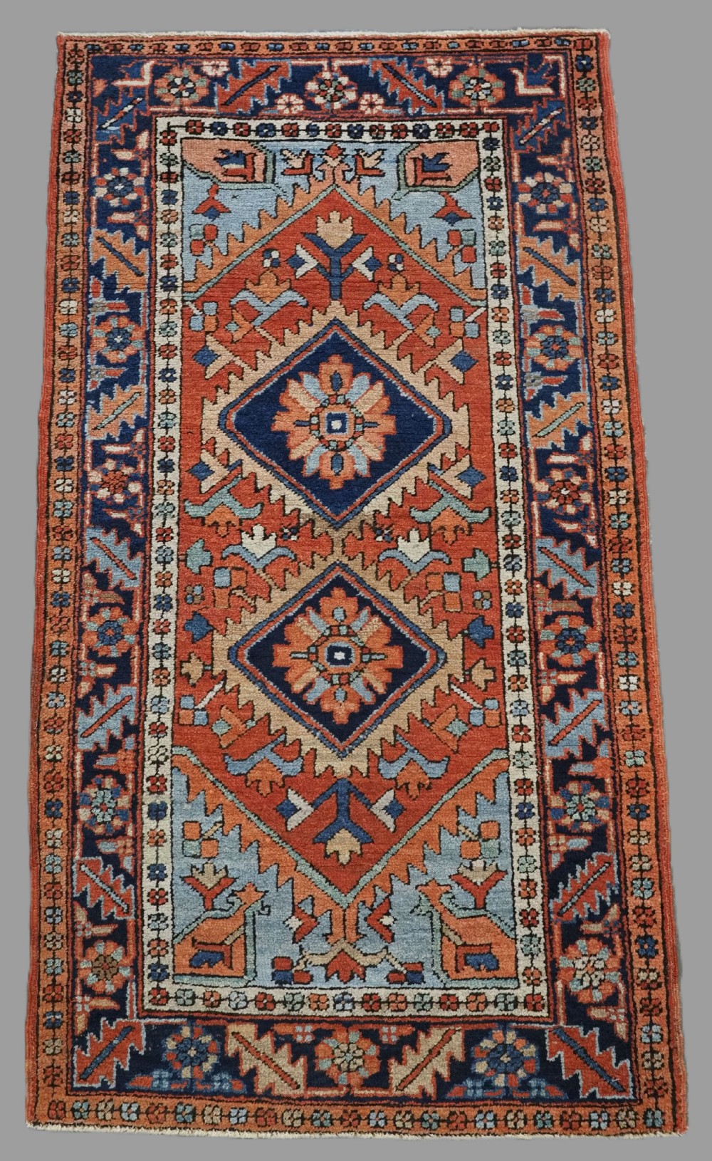 A small rectangular hand-knotted antique Persian Karaja rug featuring central diamond medallions on a terracotta field with blue geometric borders.