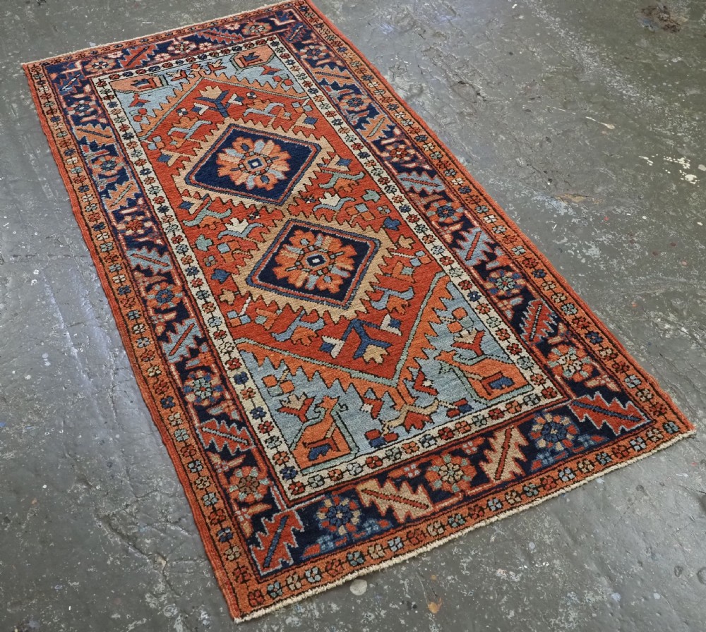 A small rectangular hand-knotted antique Persian Karaja rug featuring central diamond medallions on a terracotta field with blue geometric borders. View II