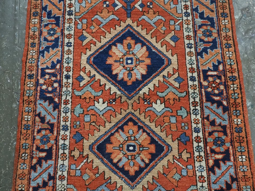 A small rectangular hand-knotted antique Persian Karaja rug featuring central diamond medallions on a terracotta field with blue geometric borders. View IV