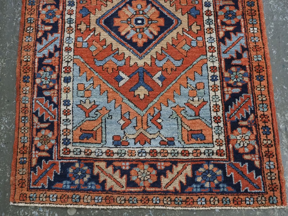 A small rectangular hand-knotted antique Persian Karaja rug featuring central diamond medallions on a terracotta field with blue geometric borders. View V