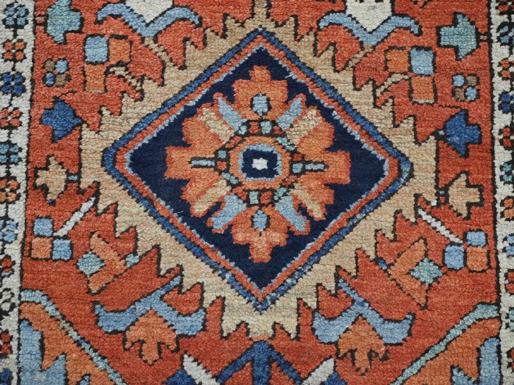A small rectangular hand-knotted antique Persian Karaja rug featuring central diamond medallions on a terracotta field with blue geometric borders. View X