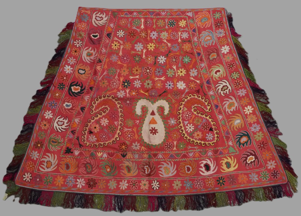 A trapezoidal Central Asian textile featuring dense multi-coloured floral, boteh, and horn embroidery on a red background with fringed edges.