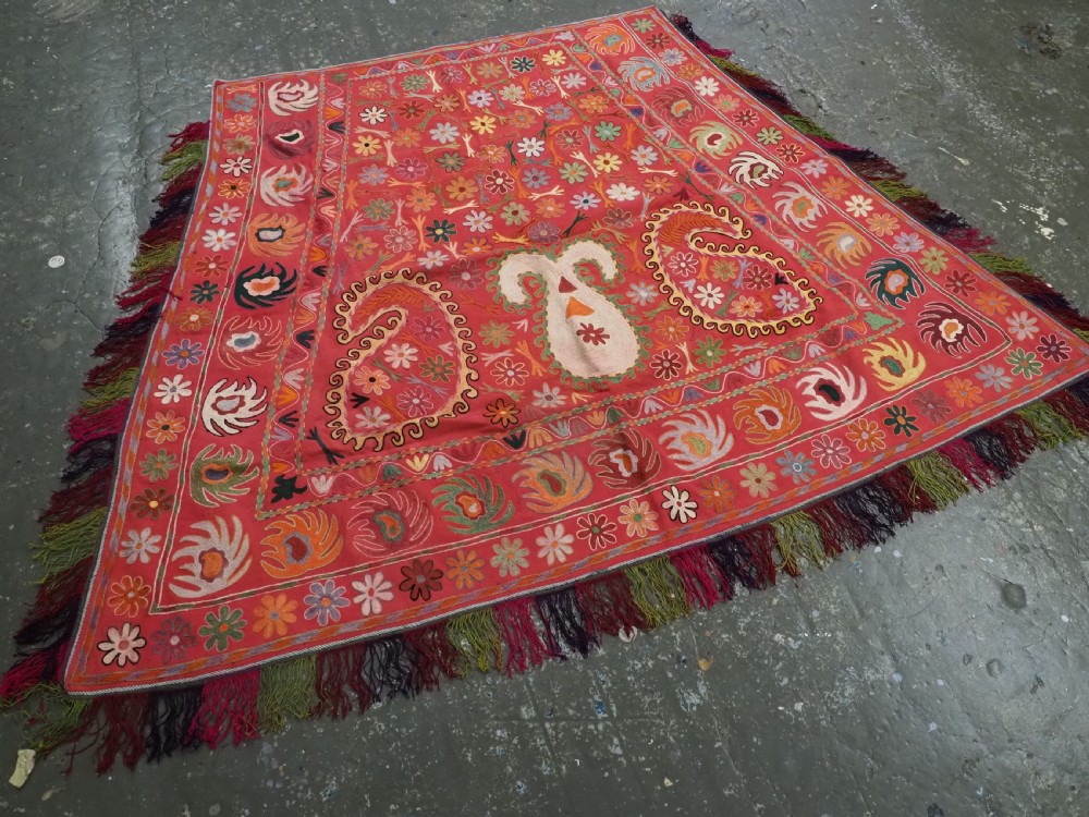 A trapezoidal Central Asian textile featuring dense multi-coloured floral, boteh, and horn embroidery on a red background with fringed edges. View II