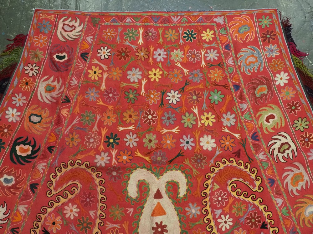 A trapezoidal Central Asian textile featuring dense multi-coloured floral, boteh, and horn embroidery on a red background with fringed edges. View III