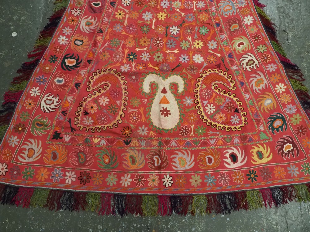 A trapezoidal Central Asian textile featuring dense multi-coloured floral, boteh, and horn embroidery on a red background with fringed edges. View IV