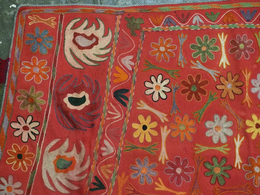 A trapezoidal Central Asian textile featuring dense multi-coloured floral, boteh, and horn embroidery on a red background with fringed edges. View V