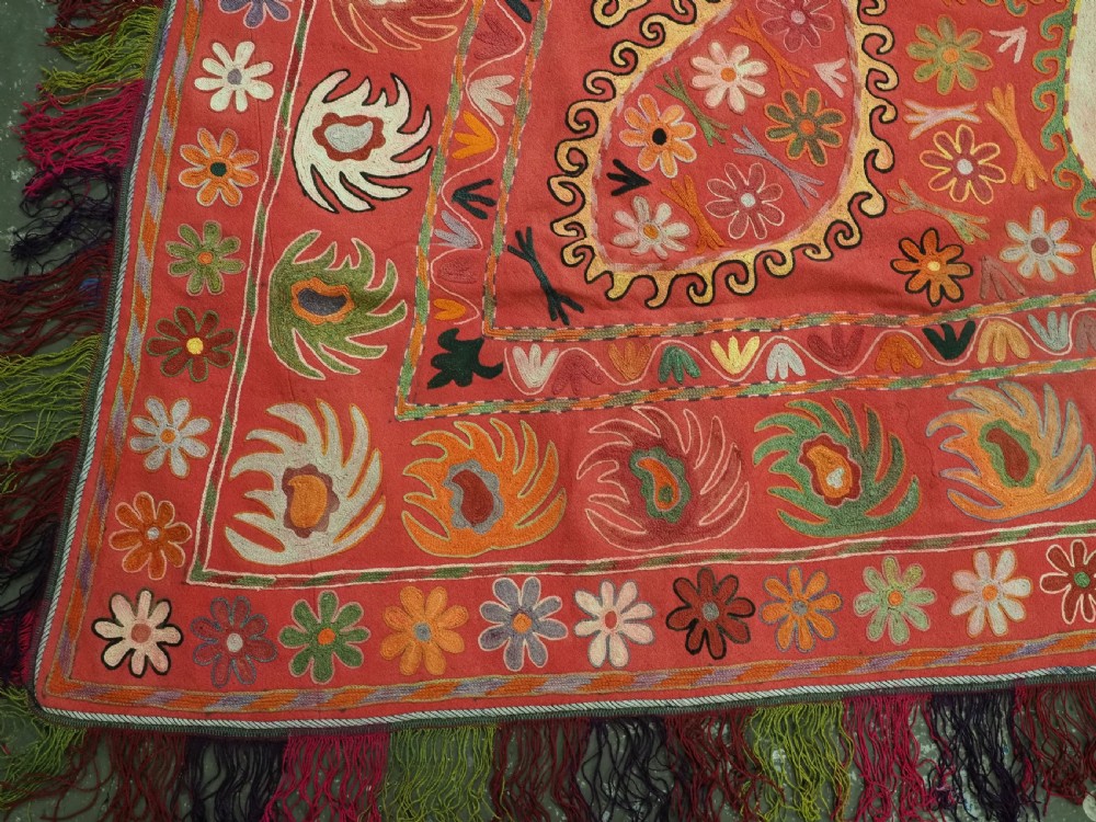 A trapezoidal Central Asian textile featuring dense multi-coloured floral, boteh, and horn embroidery on a red background with fringed edges. View X
