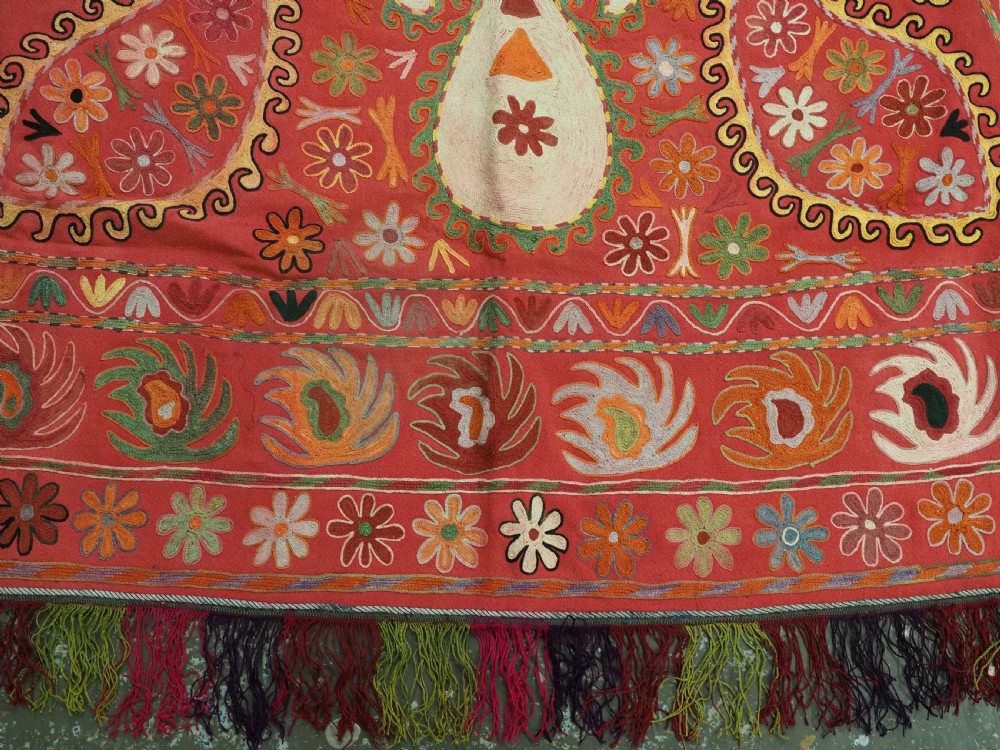 A trapezoidal Central Asian textile featuring dense multi-coloured floral, boteh, and horn embroidery on a red background with fringed edges. View XI