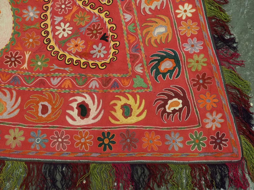 A trapezoidal Central Asian textile featuring dense multi-coloured floral, boteh, and horn embroidery on a red background with fringed edges. View XII