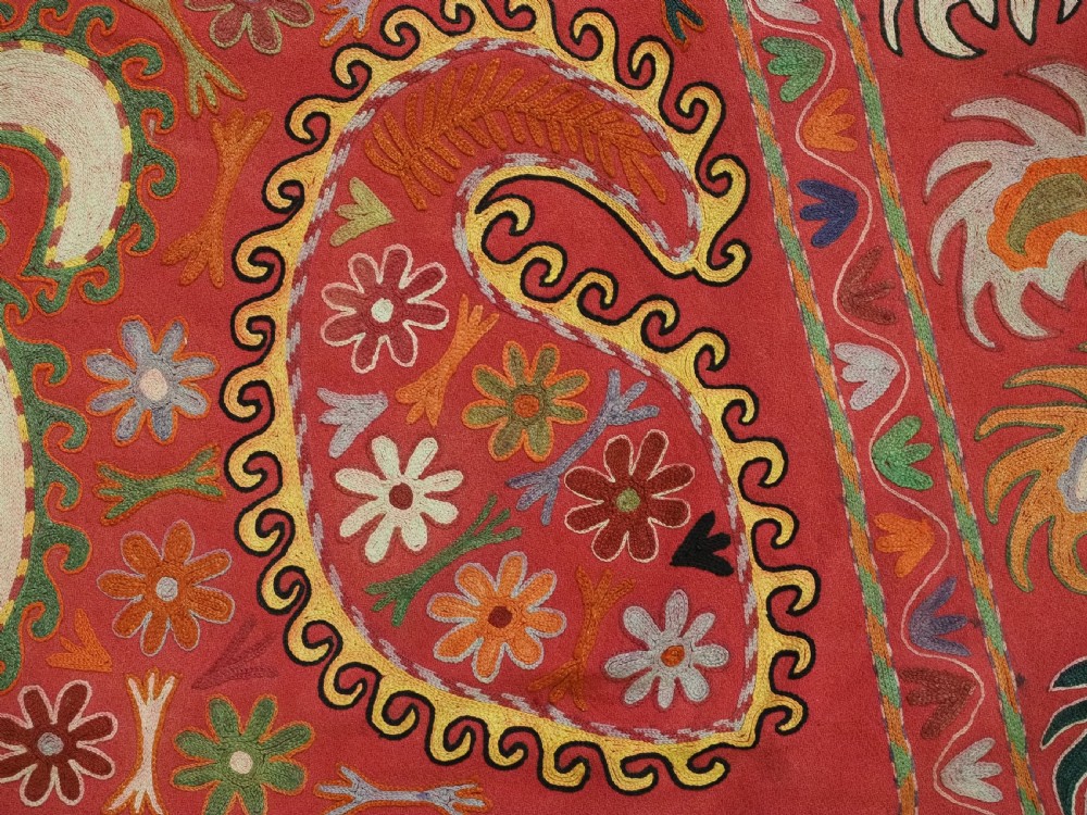 A trapezoidal Central Asian textile featuring dense multi-coloured floral, boteh, and horn embroidery on a red background with fringed edges. View XIII