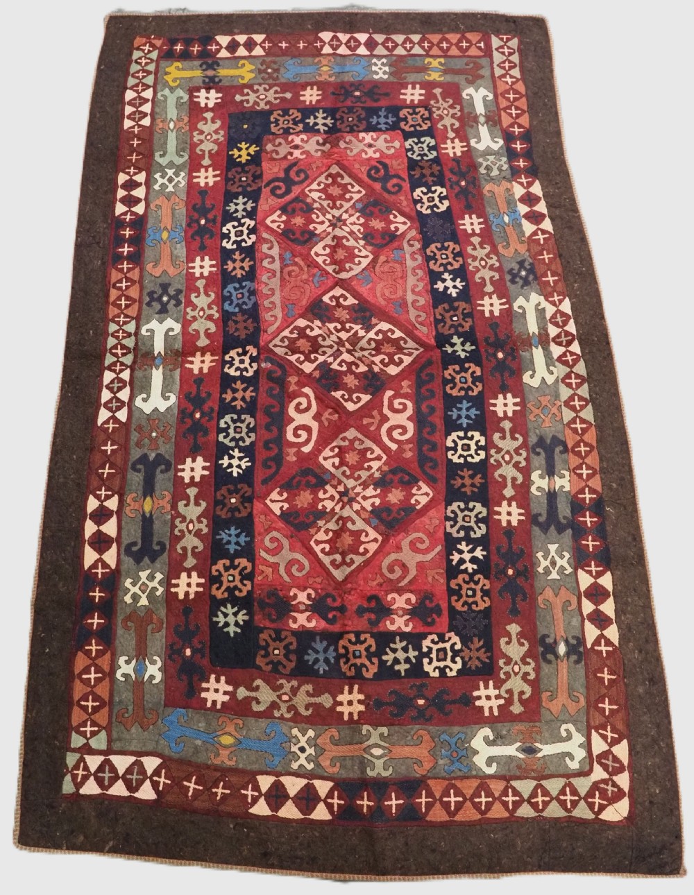 An antique Central Asian embroidered felt rug featuring a terracotta red centre with diamond medallions surrounded by multi-coloured geometric borders on dark brown felt.