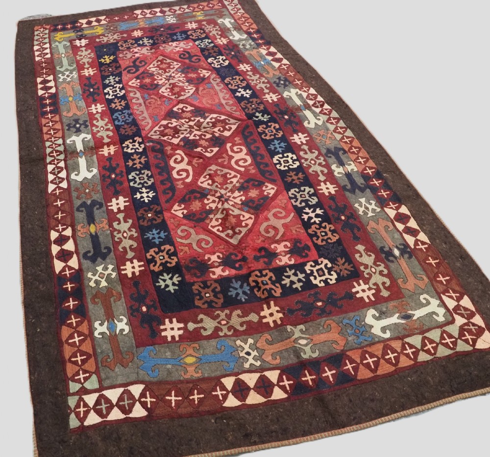 An antique Central Asian embroidered felt rug featuring a terracotta red centre with diamond medallions surrounded by multi-coloured geometric borders on dark brown felt. View II