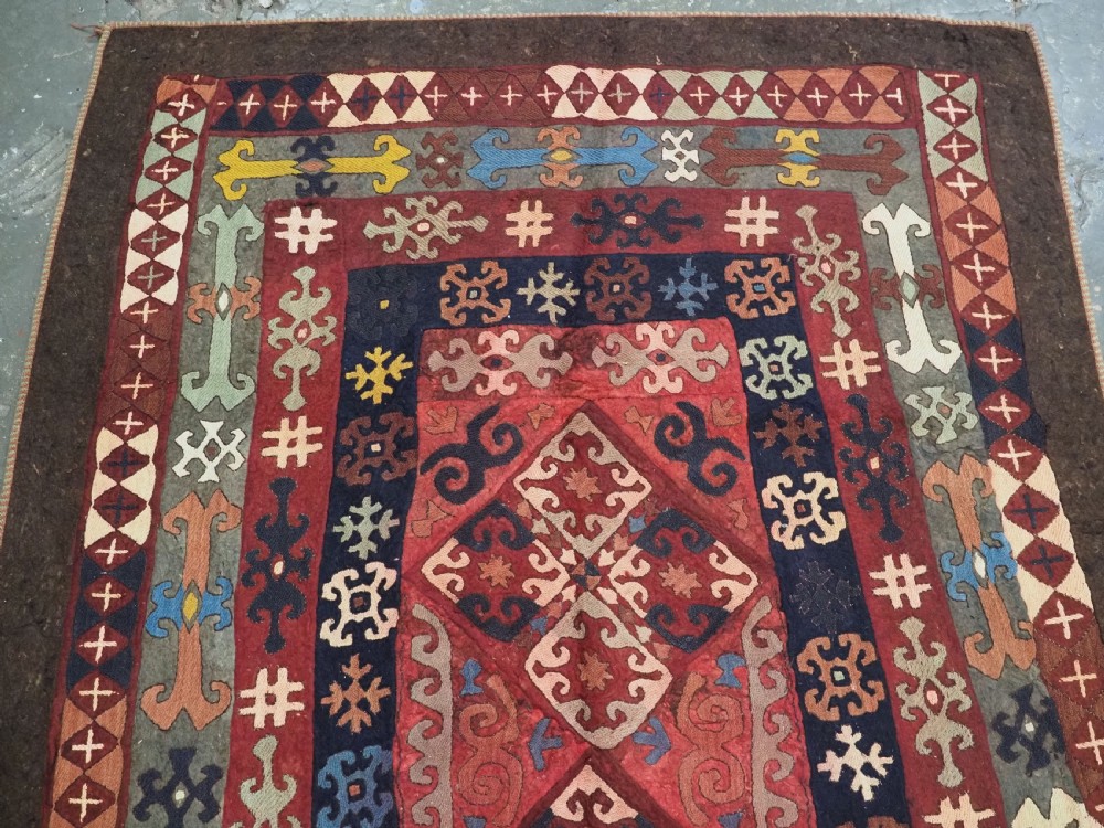 An antique Central Asian embroidered felt rug featuring a terracotta red centre with diamond medallions surrounded by multi-coloured geometric borders on dark brown felt. View III