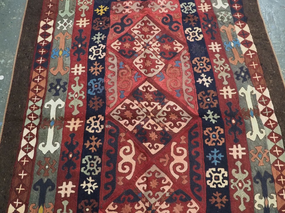 An antique Central Asian embroidered felt rug featuring a terracotta red centre with diamond medallions surrounded by multi-coloured geometric borders on dark brown felt. View IV