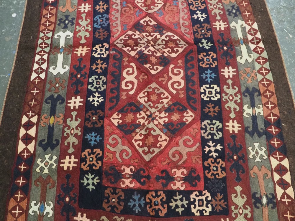 An antique Central Asian embroidered felt rug featuring a terracotta red centre with diamond medallions surrounded by multi-coloured geometric borders on dark brown felt. View V