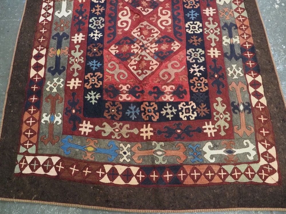An antique Central Asian embroidered felt rug featuring a terracotta red centre with diamond medallions surrounded by multi-coloured geometric borders on dark brown felt. View VI