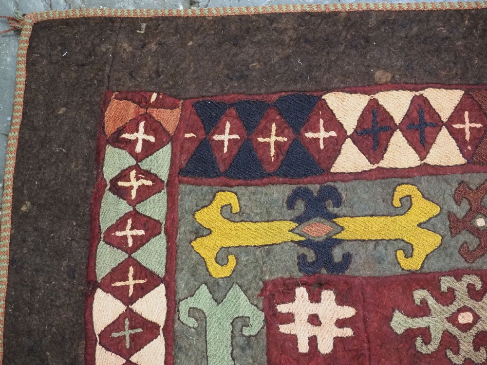 An antique Central Asian embroidered felt rug featuring a terracotta red centre with diamond medallions surrounded by multi-coloured geometric borders on dark brown felt. View VII