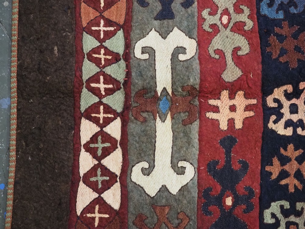 An antique Central Asian embroidered felt rug featuring a terracotta red centre with diamond medallions surrounded by multi-coloured geometric borders on dark brown felt. View VIII