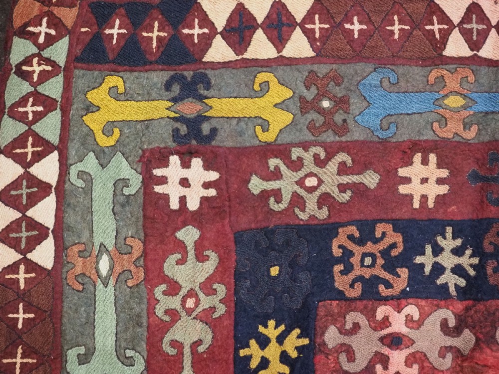 An antique Central Asian embroidered felt rug featuring a terracotta red centre with diamond medallions surrounded by multi-coloured geometric borders on dark brown felt. View IX